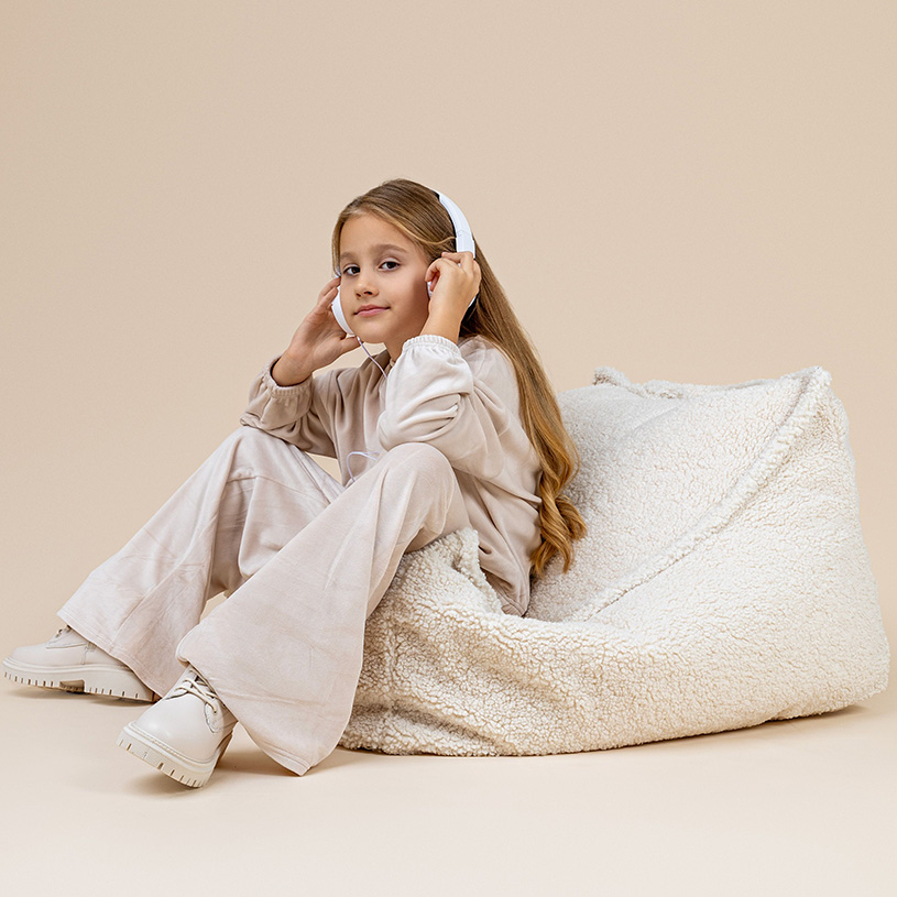 Pige i beige tøj lytter til høretelefoner, mens hun sidder på en stor, lys puf.